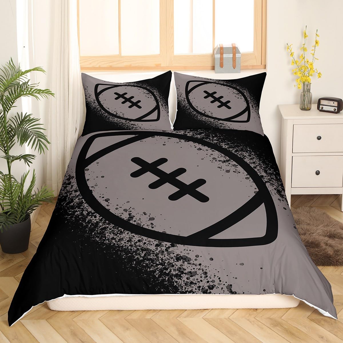 A bed dressed in a black and gray football-themed bedding set with two matching pillowcases, shown in a styled bedroom setting with a window, plant, and side table.