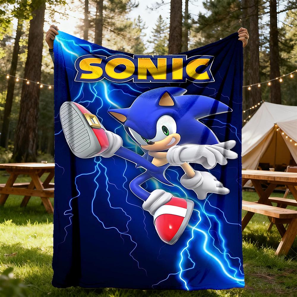 A fleece blanket featuring Sonic the Hedgehog is held up outdoors in a camp setting. The blanket is dark blue with Sonic running through blue lightning, featuring the yellow 'SONIC' logo at the top and his red and white sneakers.