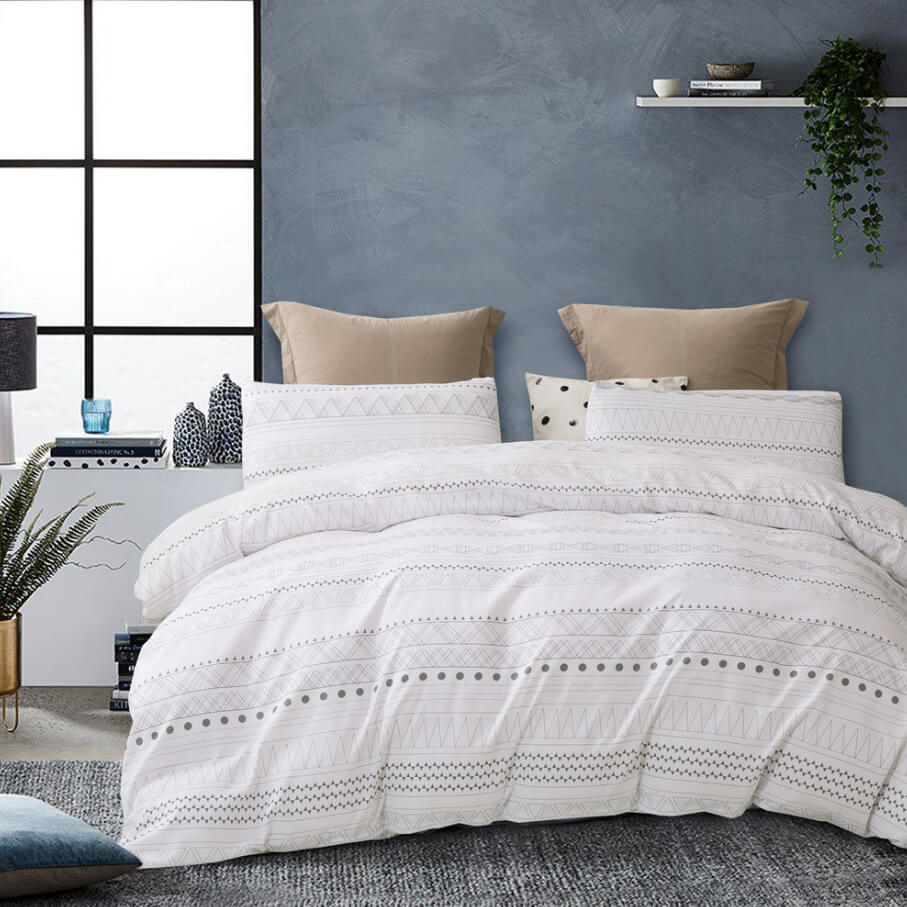 A white textured bedding set with geometric embroidered patterns, styled on a bed in a modern bedroom with a blue-gray wall, beige accent pillows, and a floating shelf.