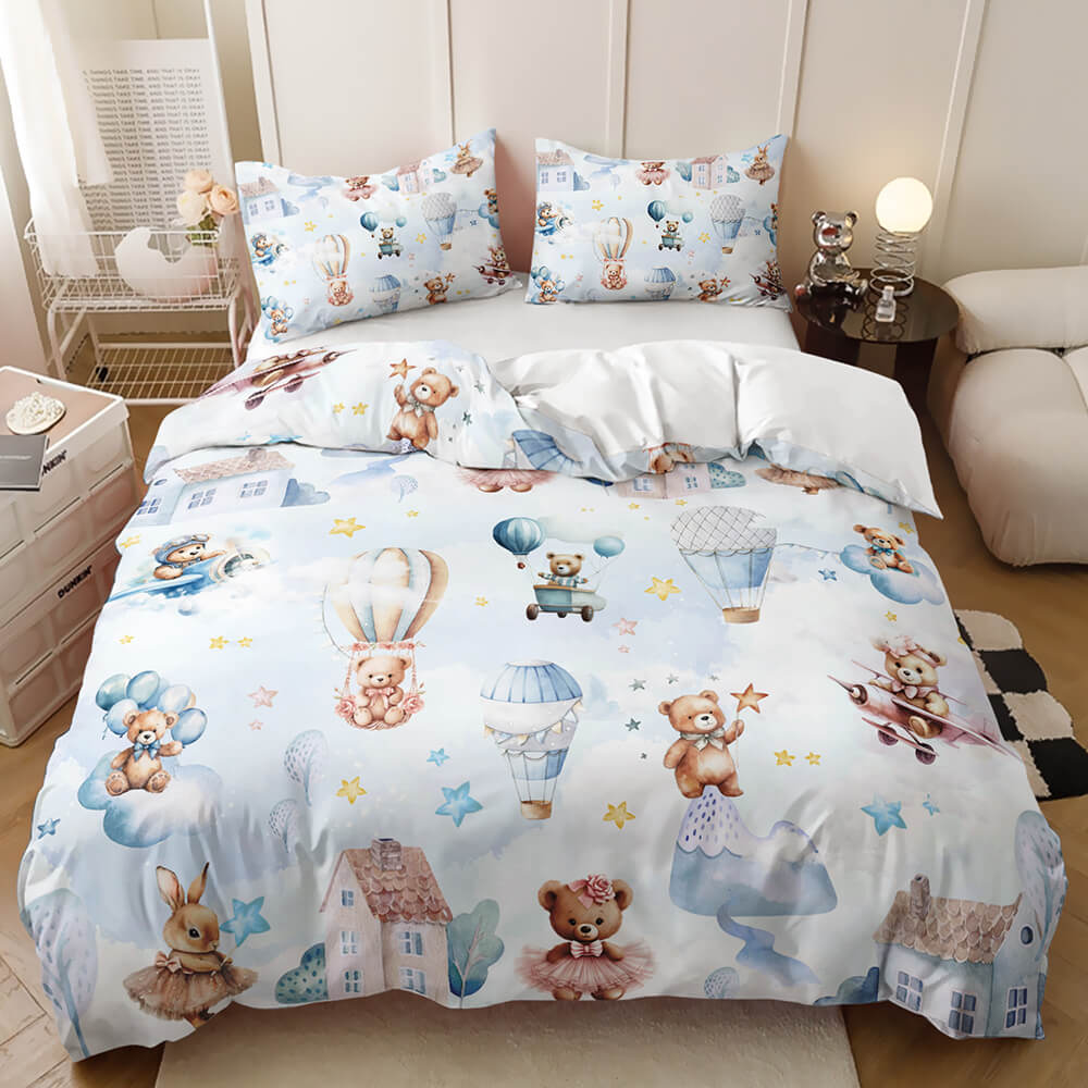 A light blue baby bedding set with a whimsical cartoon print of teddy bears, hot air balloons, stars, and small houses, styled on a bed in a bedroom setting.