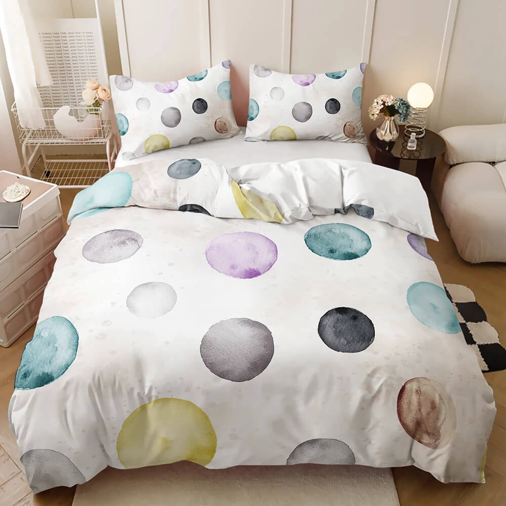 A made bed in a styled bedroom setting featuring a white duvet cover and matching pillowcases printed with large watercolor-style polka dots in pastel blue, purple, gray, yellow, and beige tones.