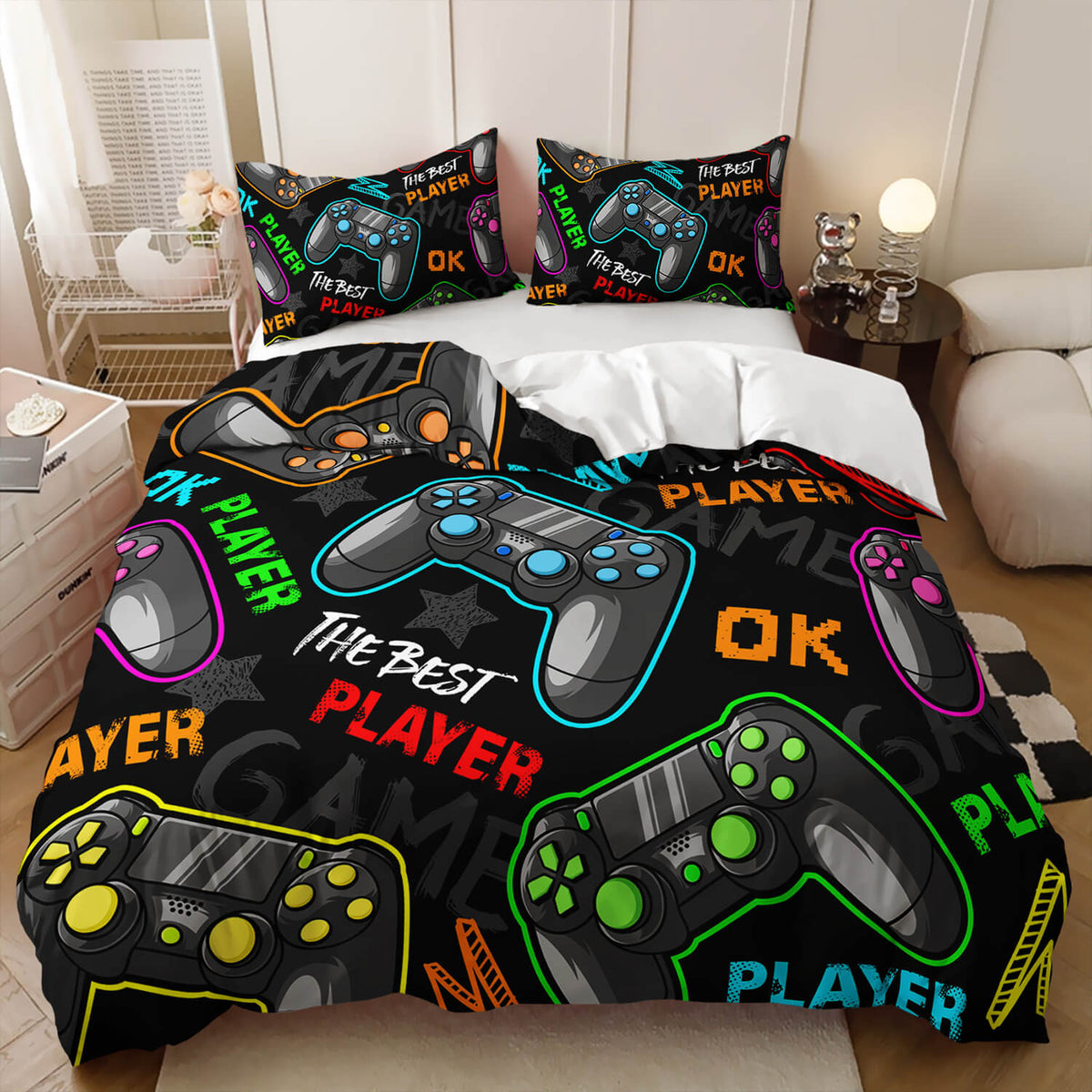 A bed made up with a black bedding set featuring large video game controller graphics and colorful neon-style text. The set includes a duvet cover and two matching pillowcases, shown in a styled bedroom with bedside tables, a lamp, and a sofa.