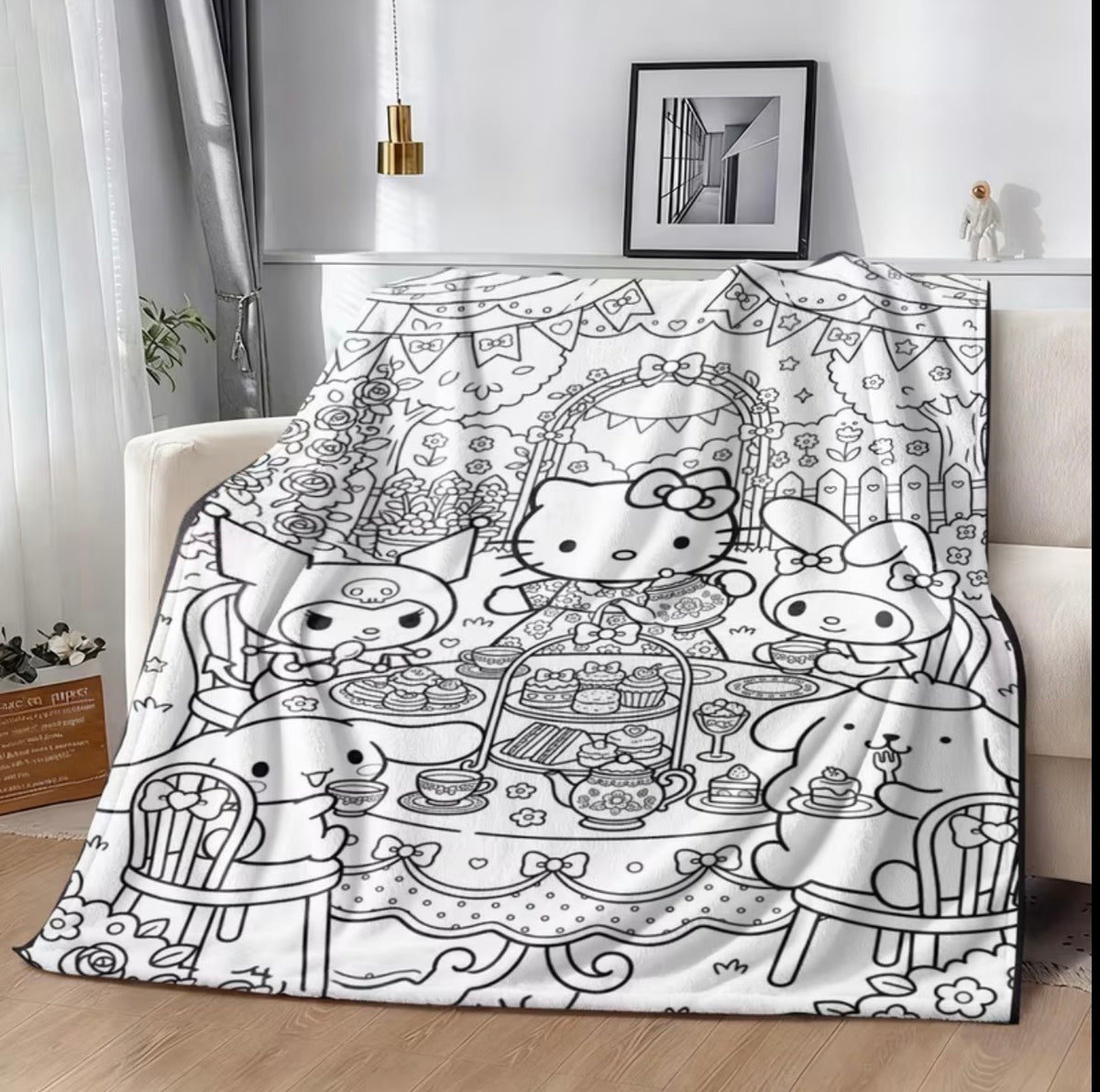 A white blanket or throw with a black line-art illustration of Hello Kitty and other Sanrio characters sitting at a tea table, draped over a beige sofa in a modern living room setting.