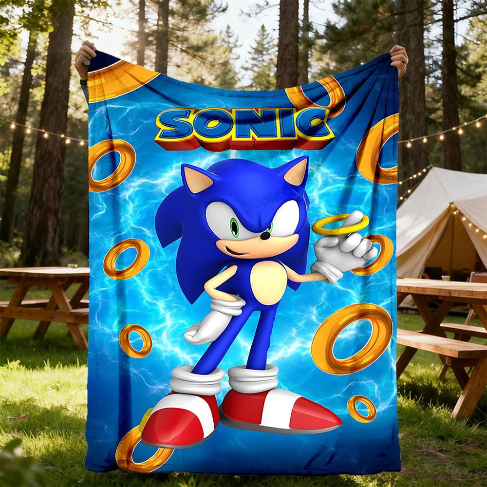 A large printed blanket or towel featuring Sonic the Hedgehog on a blue background with yellow rings and the word "SONIC" at the top, being held up outdoors by two people in a wooded campsite setting.