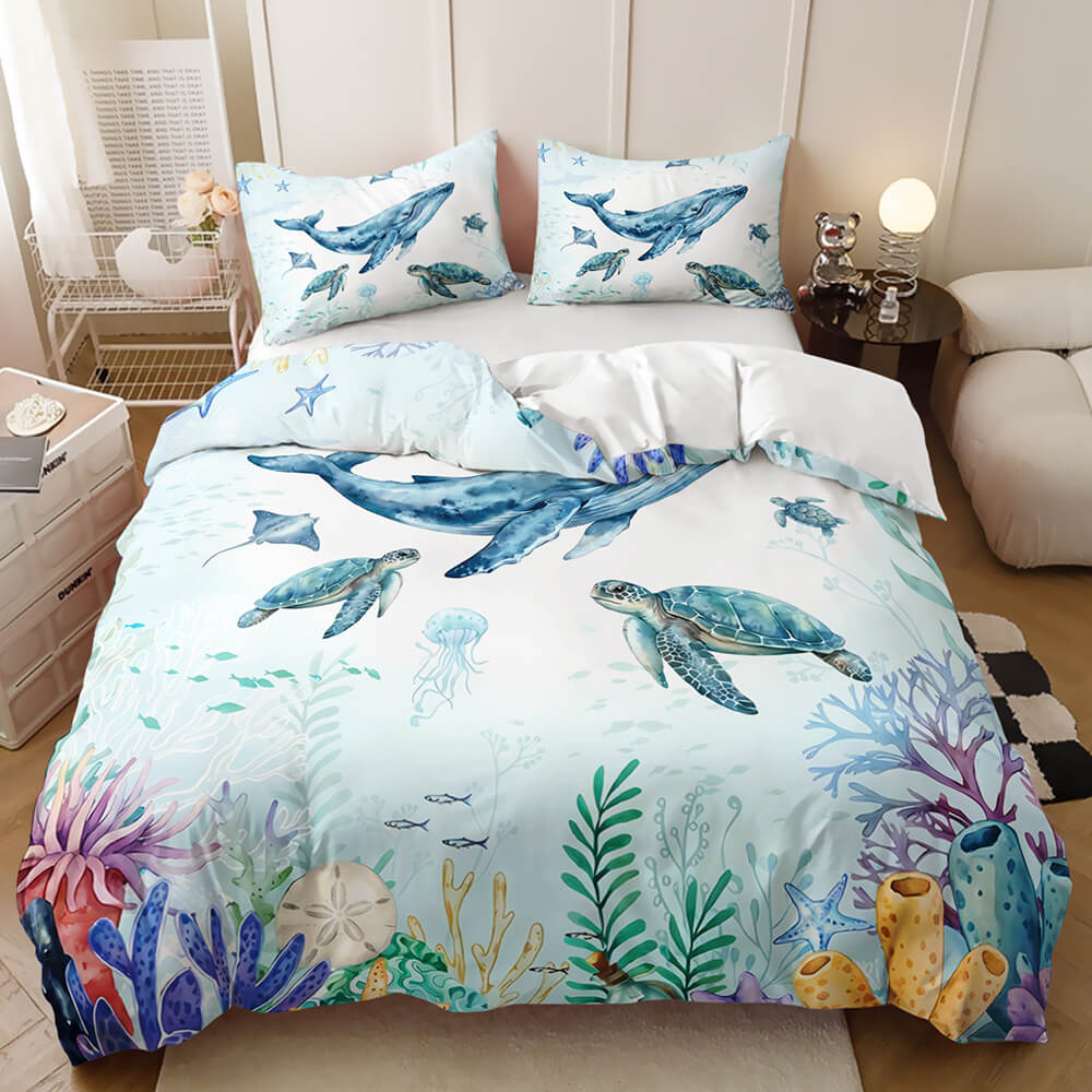 An ocean-themed bedding set featuring a duvet cover printed with blue whales, sea turtles, jellyfish, coral, and starfish on a light blue background, styled in a modern bedroom setting.