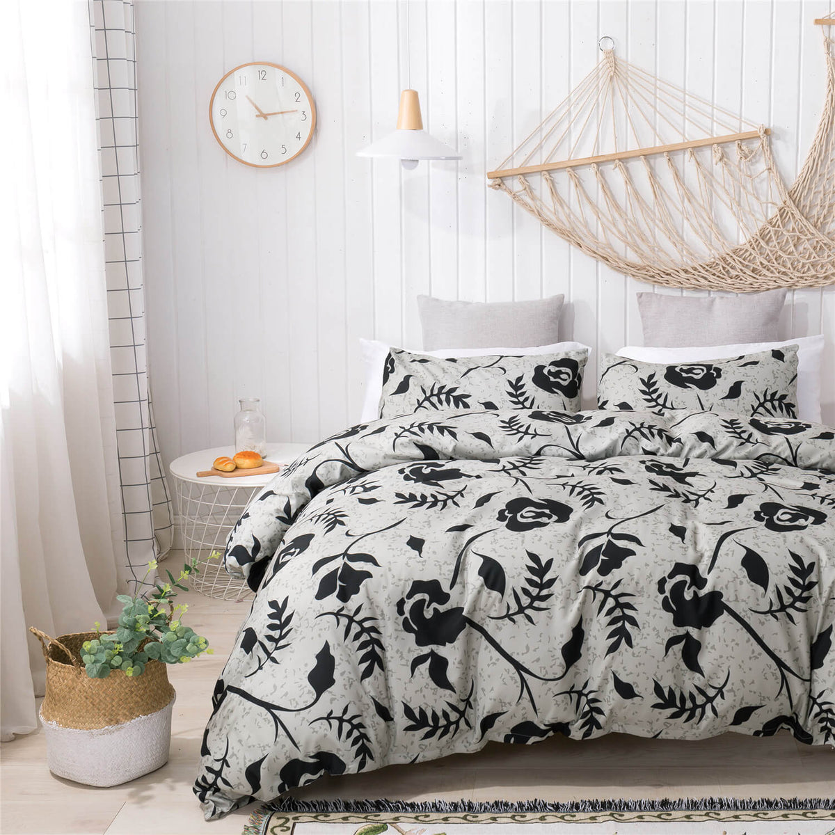 A bed dressed in a light gray duvet cover and matching pillowcases with a bold black floral and leaf pattern, styled in a bright, modern bedroom with white paneled walls, a macrame hammock, and a small side table.
