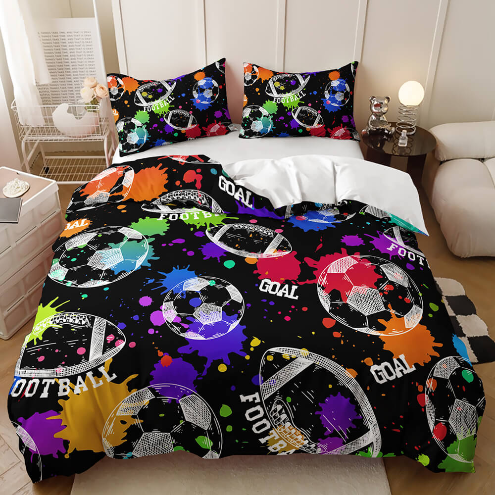 A bed dressed in a black duvet cover and matching pillowcases featuring a colorful graffiti-style football pattern with paint splatters, soccer balls, and the word 'GOAL'. The bed is styled in a bedroom setting with bedside tables, a lamp, and a sofa.